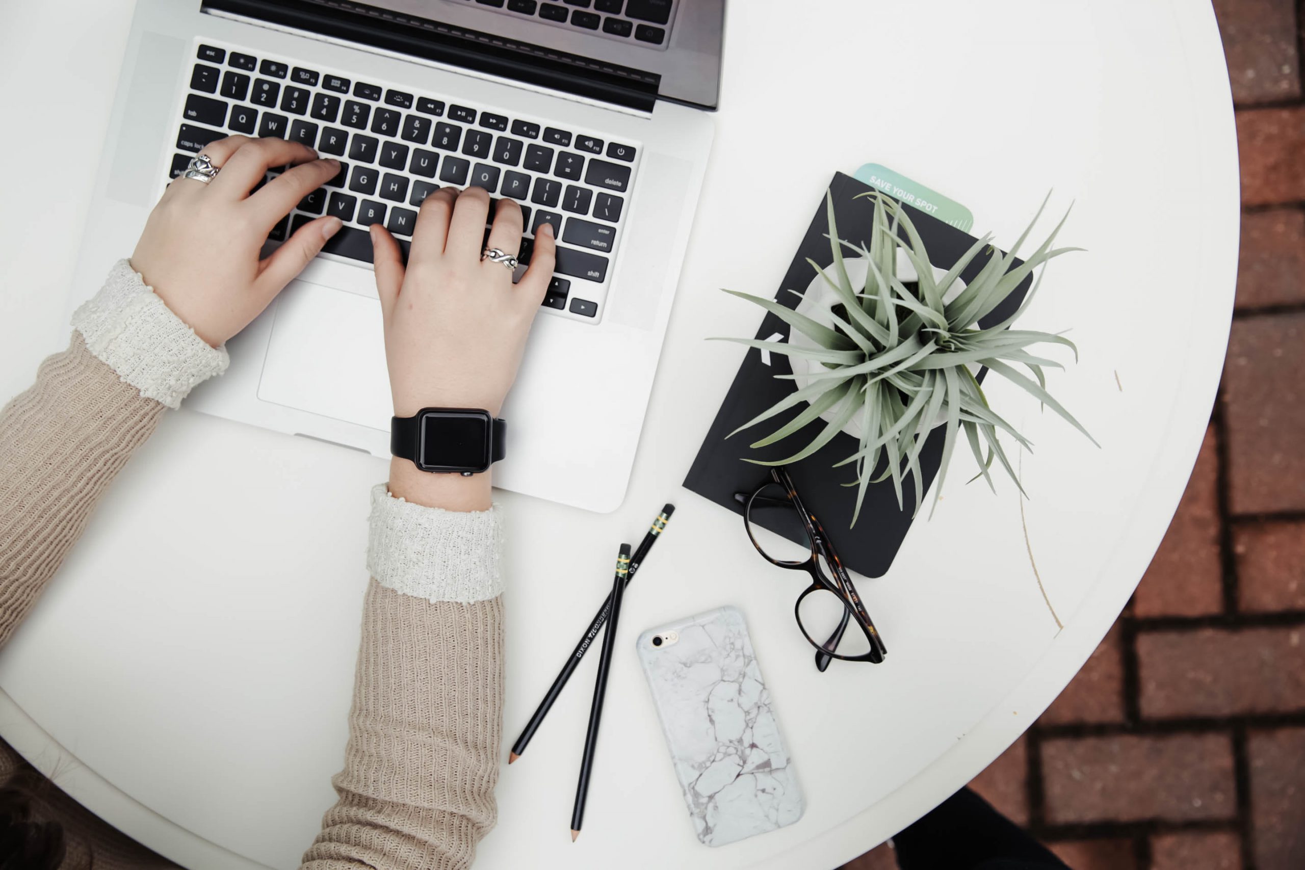 A person typing on a laptop with a plant, pencils, glasses and mobile phone next to her.